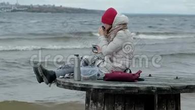 年轻女子穿着暖和的衣服坐在海边，坐在木盘管上，用<strong>热水</strong>瓶<strong>喝</strong>热茶