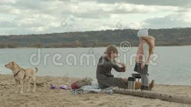 一位穿着外套的年轻女子坐在河边的海滩上，大海，野餐，从热水瓶里倒热茶，一杯棕色的