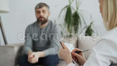 <strong>心理</strong>治疗师或<strong>心理</strong>学家在与坐在办公室沙发上的成年男子交谈时写笔记