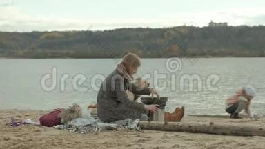一位穿着外套的年轻女子坐在河边的海滩上，大海，野餐，从<strong>热水</strong>瓶里<strong>倒</strong>热茶，一杯棕色的