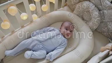 出生的小男孩躺在婴儿床上看着相机，微笑着。 可爱的小婴儿躺在家里的婴儿床上。 小男孩