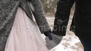 特写新郎牵着新娘的手在冬天下雪时在松林散步的视频。 下雪