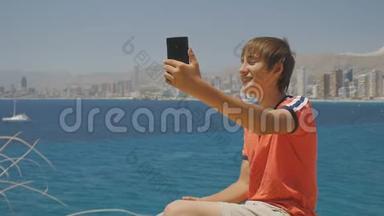 英俊的微笑少年在海上海岸和摩天大楼天际线背景下制作智能手机视频通话的肖像。