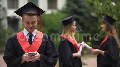 男毕业生用手机打字，用相机微笑，毕业
