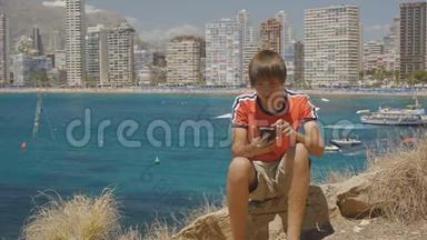 15岁的男孩坐在海边的石头上，在摩天大楼的天际线背景下用智能手机输入信息。