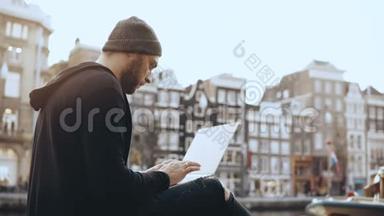 4K创意人坐在街上拿着笔记本电脑。 工作移动办公室。 阿姆斯特丹老城区。 大气城市景观。