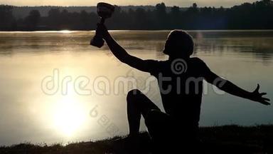 金发碧眼的人坐在湖岸上，看着一只盛满水的冠军碗