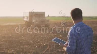英俊的农民与平板站在联合收割机的背景。 农民使用现代技术触摸平板电脑