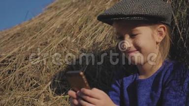 年轻可爱的女孩带着帽子玩得很开心，坐在干草堆里用电话。 坐在干草上的小女孩。