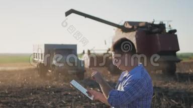 英俊的农民与平板站在联合收割机的背景。 农民使用现代技术触摸平板电脑