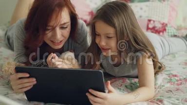 一位微笑可爱的女孩和年轻的母亲穿<strong>着</strong>睡衣，<strong>躺</strong>在床上笑<strong>着看着</strong>数码平板电脑。