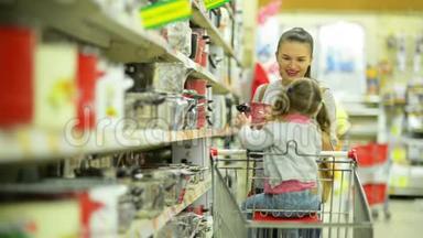 超市年轻幸福家庭肖像。 可爱的小女孩坐在购物车里，年轻的女人选择