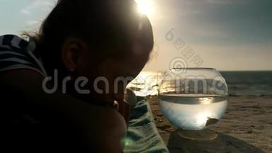 小女孩躺在海滩上，看着夕阳下鱼缸里的小鱼。