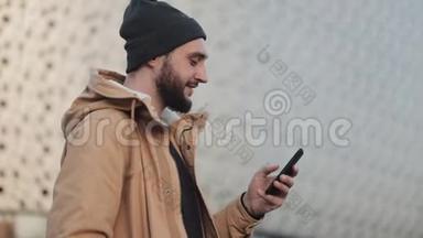 快乐的年轻胡须男用智能手机在购物中心附近的街道上行走。 他穿着一件秋装