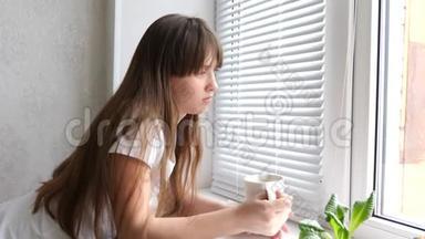 美丽悲伤的女孩望着窗外喝茶或咖啡。