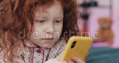 有趣的红头发小女孩与黄色智能<strong>手机躺</strong>在床上和使用智能<strong>手机</strong>。 沟通，播放，APP..