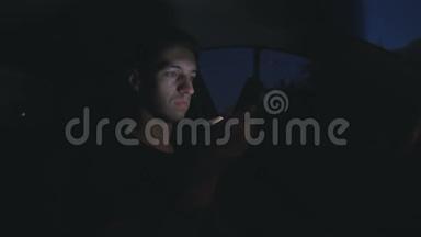 一个年轻人在夜间坐在马路上的车里用智能手机