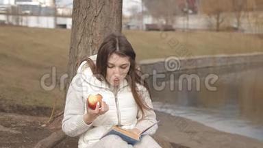美丽的<strong>女孩</strong>在公园里的一棵树下吃一个红苹果，在<strong>水边</strong>看书