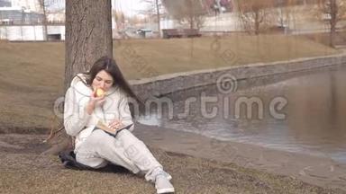美丽的女孩在公园里的一棵树下吃一个红苹果，在水边看书