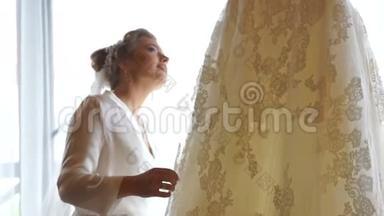 美丽的美丽的新娘，带着美丽的微笑，正在享受她挂在衣架上的婚纱。