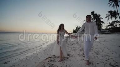 在海边日落时新婚快乐。 他们赤着脚走在沙滩上，手牵着手，看着每一个人