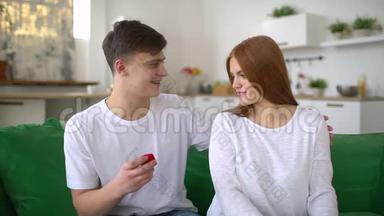 年轻男子向坐在沙发上的戴着<strong>红礼盒</strong>戒指的吃惊女人求婚，令幸福的女人吃惊