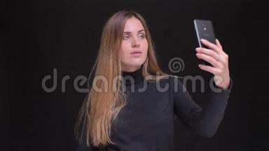 年轻的白种人长发金发金发女孩在黑色背景下使用智能手机制作自拍照片的肖像。