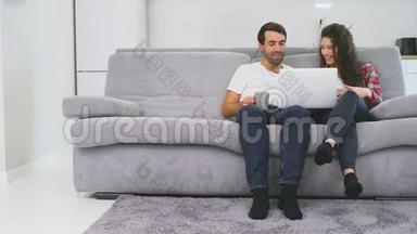 幸福的男人和幸福的男人在家里一起坐在沙发上，微笑着的年轻夫妇用电脑享受早晨的咖啡