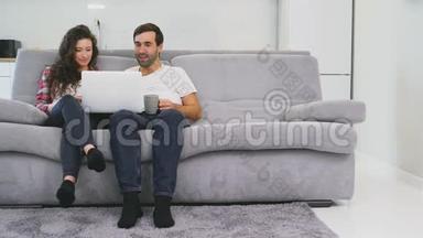 <strong>幸福</strong>的男人和<strong>幸福</strong>的男人在家里一起坐在沙发上，微笑着的年轻夫妇用电脑享受早晨的咖啡