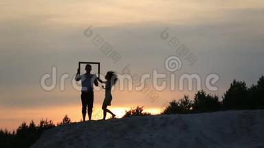 在夕阳的背景下，一对滑稽有趣的浪漫情侣抱着空框接吻