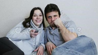 两个人躺在沙发上看电视。 女人有兴趣地切换频道和观看，男人很无聊。