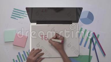 女人在笔记本电脑上的一张纸上写TEAMWORK。 带有图表的业务桌面。
