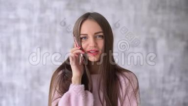 穿着粉色衬衫的快乐美女在智能手机前聊天，她微笑着看着灰色背景下的相机