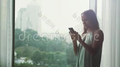 年轻漂亮的黑发女商人用手机站在全景窗口与城市景色。 4k，3840x2160