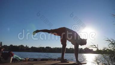 年轻运动员练习瑜伽动作的剪影和在湖边木制码头的位置。 运动员训练