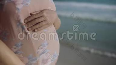 年轻的孕妇站在蓝色海洋附近的海滩上呼吸，靠近手和腹部。 暑假假期