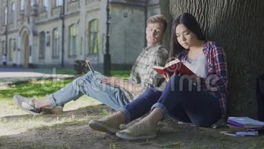 一个手提电脑的男人坐在树下看着女孩看书