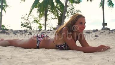 微笑的女人躺在沙滩上，在暑假的时候晒日光浴。 快乐的女人在沙滩上晒太阳