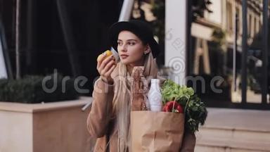 年轻漂亮的女人穿着时尚的外套站在街上，拿着一包产品。 她拿着黄色的
