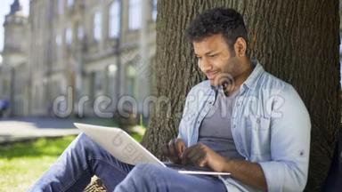 跨国青年<strong>坐在树下</strong>，在笔记本电脑上打字，微笑着聊天