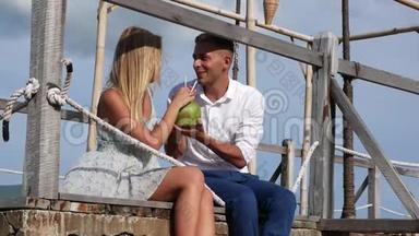 年轻的幸福夫妇坐在木墩上，笑着喝着蓝天背景上的椰子。 浪漫浪漫