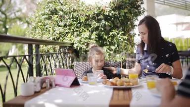 一家人在户外咖啡馆吃早餐。 可爱的女孩和妈妈在露台上喝新鲜果汁和吃牛角面包