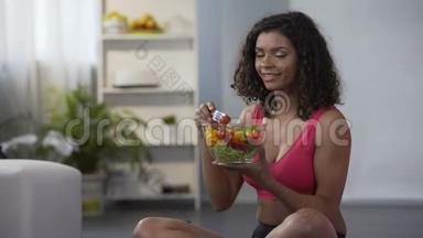 年轻健康的女人吃蔬菜沙拉，坐在地板上穿着健身房的衣服，节食