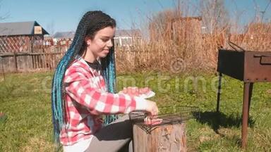 年轻的女人把切子放在烤架上。 她穿着<strong>格子粉色</strong>衬衫，有蓝色的辫子。