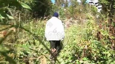 一个人在阳光明媚的夏日，在高高的绿草上走在森林里。 录像。 戴白色夹克和帽子的领班