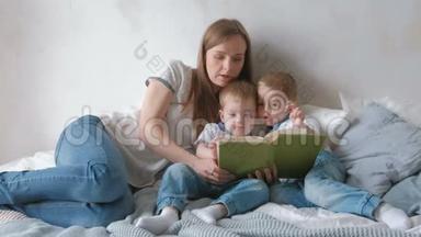 家庭妈妈和两个双胞胎兄弟蹒跚学步的孩子在床上看书。 家庭阅读时间。