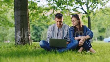年轻可爱的一对享受笔记本电脑。 <strong>坐在</strong>公园的草地上，<strong>树下</strong>