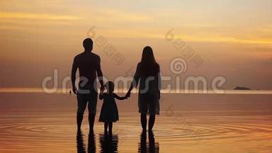 在令人惊叹的金色日落中，幸福的年轻家庭沿着海岸散步的剪影。 家庭假日。 慢动作