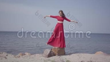 穿着红色长裙，手舞足蹈的娇艳女子举起手在蓝色海水的壮观景色附近的大石头上