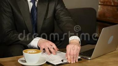 咖啡馆里的商人用商业文件工作。 男人坐在咖啡馆的桌子旁，笔记本电脑和咖啡躺在桌子上。